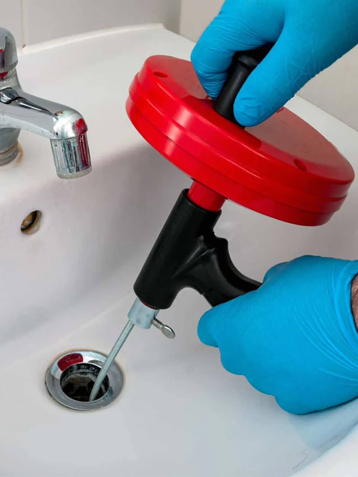 Hand-cranked drum auger being fed into a bathroom sink waste, with blue gloves and a chrome tap in shot