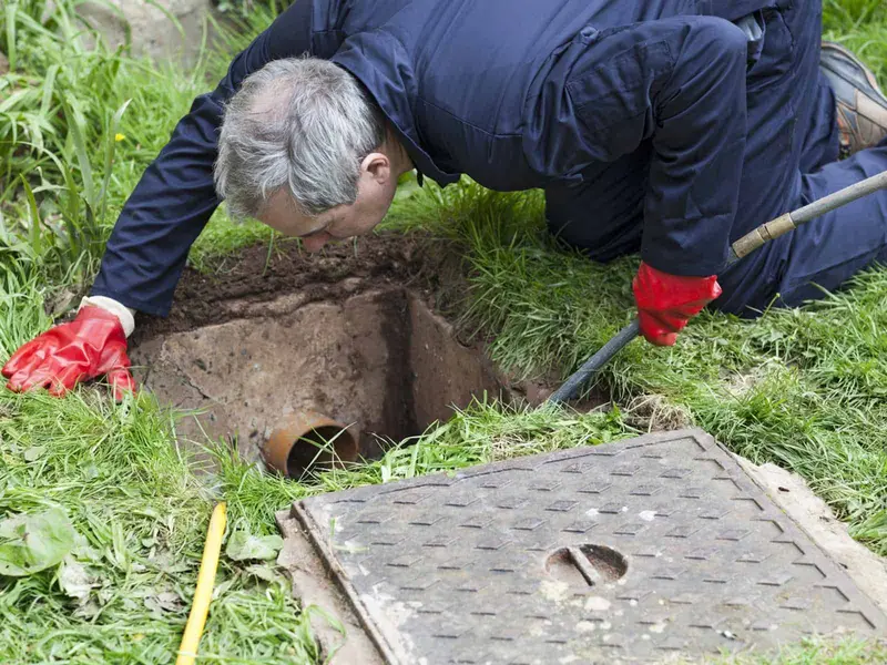 Engineer kneeling on the grass to look into an excavated chamber with a clay pipe, the cast-iron cover lifted beside it