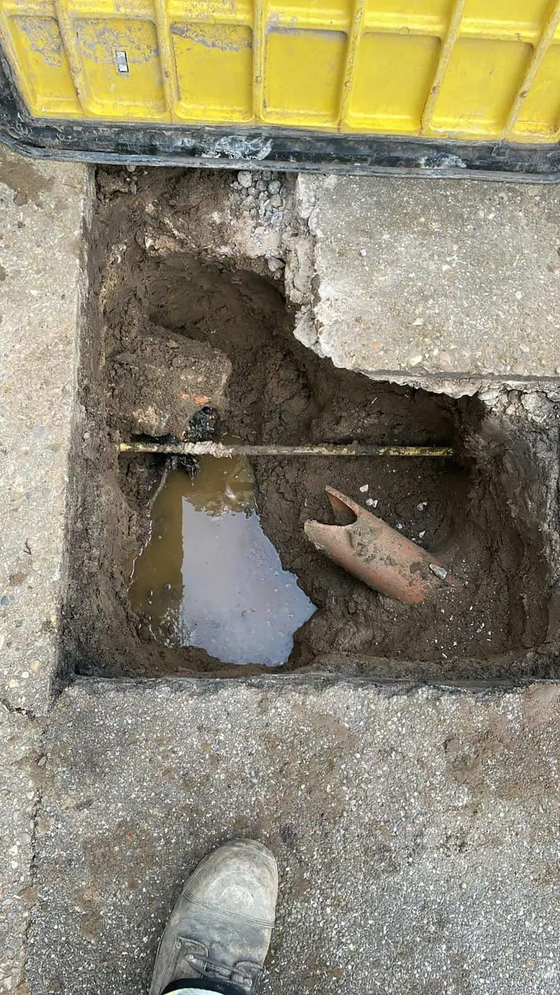 Broken clay drain pipe and standing water in a small excavation under a lifted road plate at a house in Chaddesden