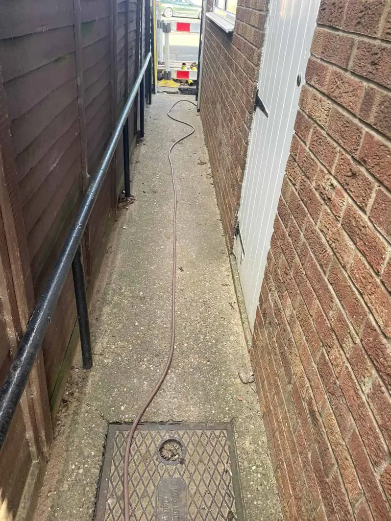 Thin cable run down a narrow side passage to a drain cover with a small access hole, at a house in Chaddesden
