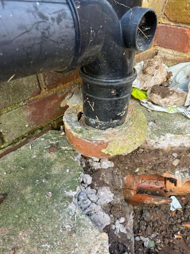 Black soil pipe dropping into a clay gully collar at the foot of a wall, with the ground beside it dug out, at a house in Alvaston